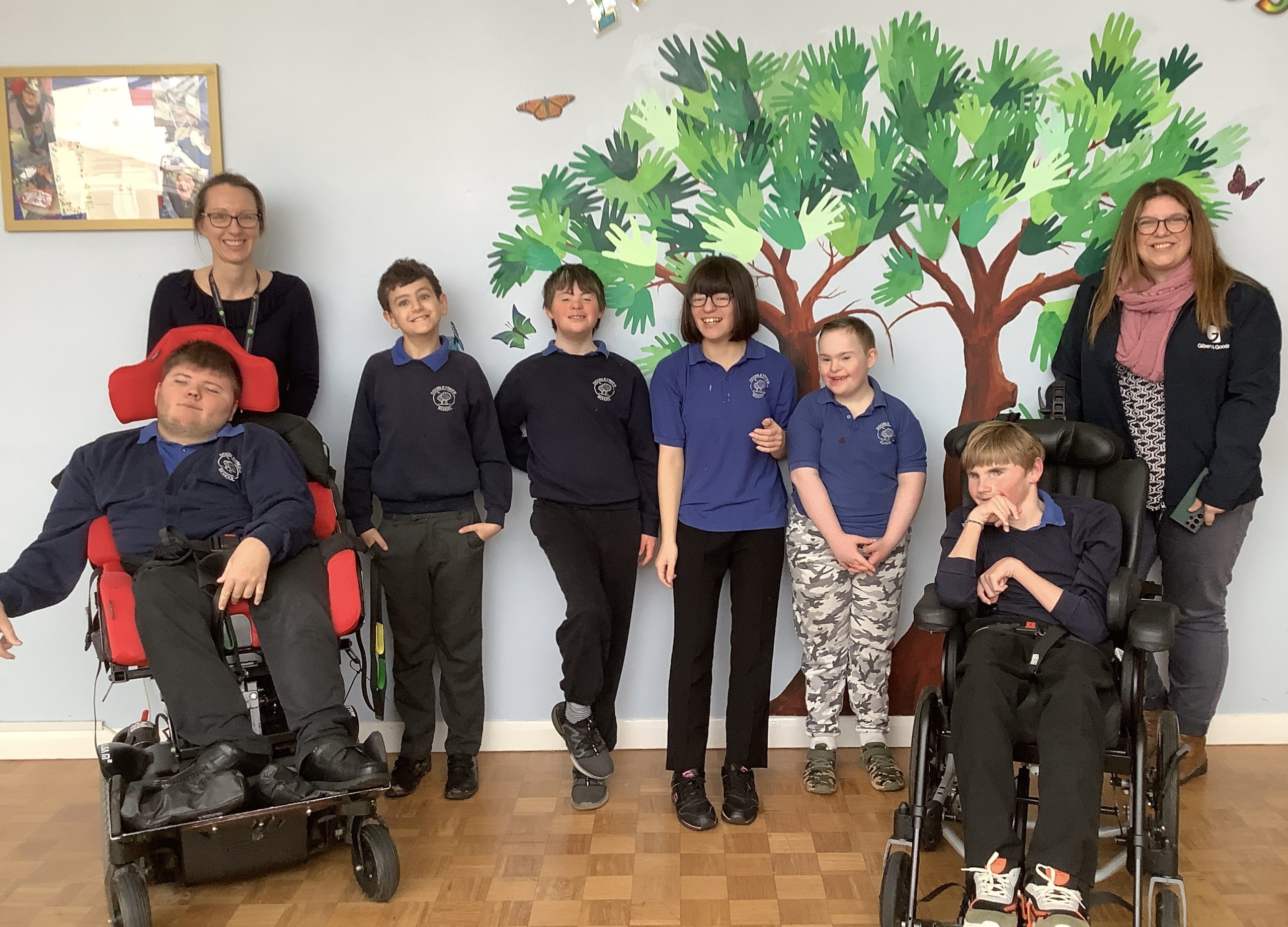 A Gilbert & Goode colleague stands in front of children’s artwork at a school, smiling alongside some of the children, following a donation to Doubletrees from Gilbert & Goode.