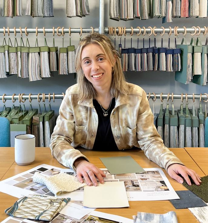 Millie, an interior designer from Clayton and Co, sits at a desk covered with various material samples, thoughtfully examining a fabric swatch.